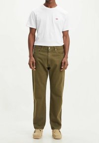 Hombre que lleva una camiseta blanca de Levi's, pantalones vaqueros verde oliva y zapatos de mocasín beige, de pie contra un fondo liso.
