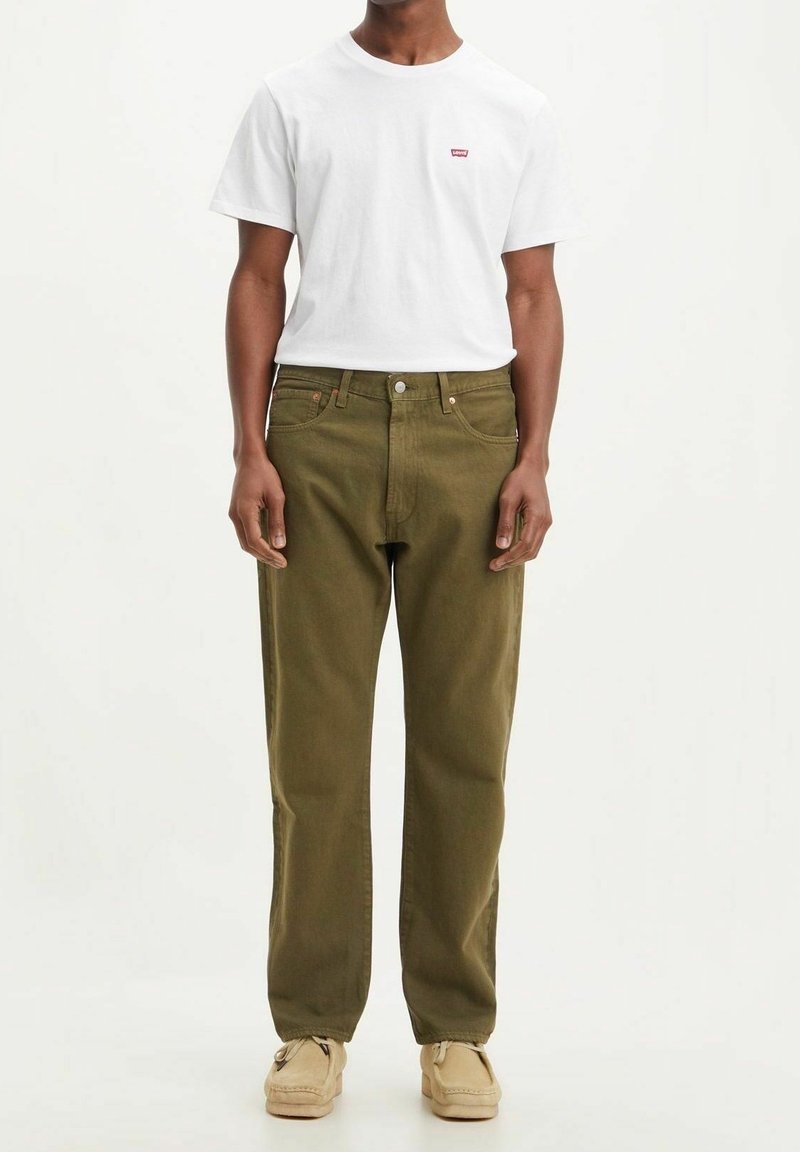 Hombre que lleva una camiseta blanca de Levi's, pantalones vaqueros verde oliva y zapatos de mocasín beige, de pie contra un fondo liso.