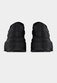 Αθλητικά παπούτσια με μαύρη χοντρή σόλα, ορατά από πίσω, που παρουσιάζουν υφασμένες καουτσούκ εξωτερικές σόλες και λείες δερμάτινες επάνω επιφάνειες με ραφές.