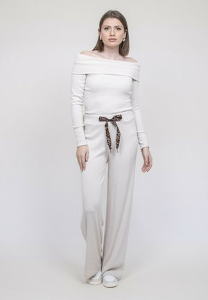 Femme debout, portant un haut blanc côtelé à épaules dénudées et un pantalon large assorti avec un cordon de serrage imprimé léopard et des baskets blanches.