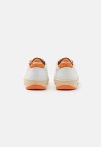 Baskets blanches avec des accents orange, tige en cuir lisse, languette arrière contrastée et semelle texturée couleur crème. Design simple avec un minimum de marque.
