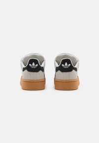 Zapatillas de ante gris con una suela de goma beige, acentos negros en el talón y un detalle de logo blanco en la parte posterior. Forma redondeada.