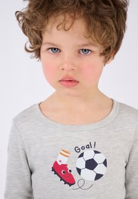 Maglietta grigia a maniche lunghe con una grafica colorata a tema calcistico, che presenta una scarpa rossa, un pallone da calcio nero e il testo giocoso "Goal!".