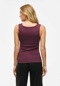 Donna con capelli biondi che indossa un top senza maniche di colore viola scuro e pantaloni neri, in piedi con le spalle rivolte verso l'osservatore su uno sfondo neutro.