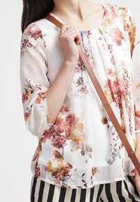 Blusa a stampa floreale in tessuto trasparente, con sfumature di fiori rosa e gialli, scollatura rotonda e maniche a tre quarti. Cintura marrone sulla spalla.