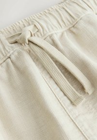 Taille élastique avec un cordon en tissu noué sur un pantalon texturé de couleur crème.