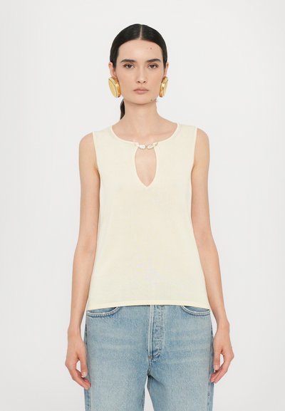 Haut sans manches jaune avec un décolleté en trou de serrure et un fermoir décoratif. Porté avec un jean en denim bleu clair. Texture lisse et design ajusté.