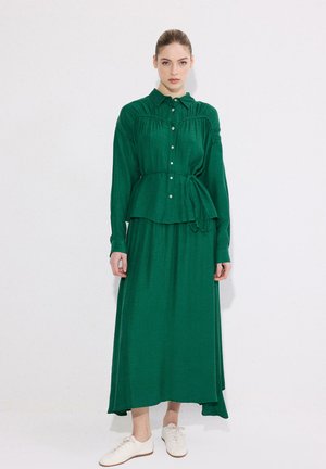 Femme debout portant un chemisier vert à manches longues avec ceinture à nouer et une jupe longue assortie, ainsi que des baskets blanches, sur fond blanc.