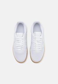 Paire de baskets blanches propres avec semelles couleur gomme, vues de dessus sur un fond gris clair.
