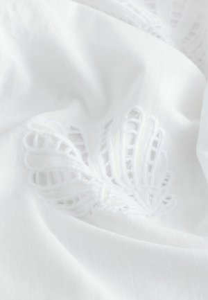 Balts audums ar fakturētām, izšūtām lapu rakstiem. Dizainā ir smalki izgriezumi, kas uzsver mīksto, gludo tekstūru.