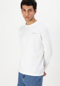 Jeune homme debout les mains derrière le dos, portant une chemise blanche à manches longues Tommy Jeans et un jean bleu, expression neutre, fond blanc.