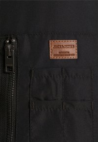 Czarna materiałowa kurtka z zamkiem z przodu, brązową skórzaną naszkliwo z napisem "JACK & JONES" oraz dwoma przednimi kieszeniami nad detalem szwów.
