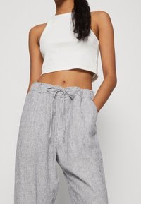 Kvinna iförd en ärmlös vit crop top och högmidjade ljusgrå randiga byxor med dragsko, står med händerna i fickorna.