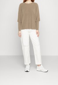 Beige geribbelde trui met een losse pasvorm en ronde halslijn, gecombineerd met witte rechte broek en witte sportschoenen.