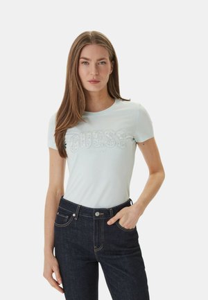 Donna con capelli castani lunghi che indossa una maglietta azzurro chiaro Guess e jeans blu scuro, con una mano in tasca.