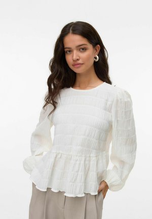 Witte peplum blouse van een textuur met een kreukelig materiaal, lange pofmouwen en een ronde halslijn. Gecombineerd met lichtgekleurde broeken.