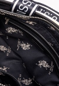 Czarny wnętrze torby z powtarzającym się, jasnym wzorem logo U.S. Polo Assn., z zamknięciem na zamek błyskawiczny i gładką teksturą materiału.