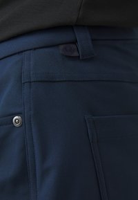 Tamnoplave hlače imaju glatku teksturu s bočnom džepom, detaljem na dugmetu i suptilnim logotipom na pojasu.