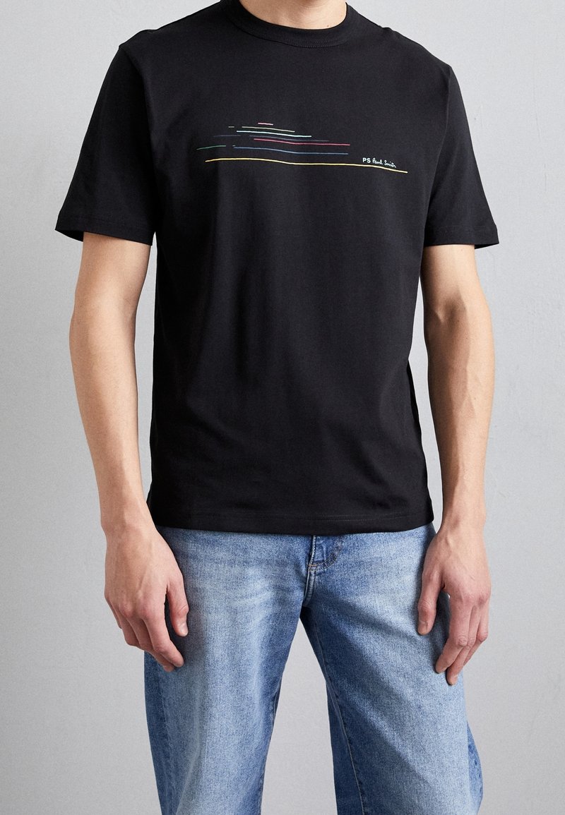 Homme portant un t-shirt noir à manches courtes avec des lignes horizontales multicolores et le texte « PS Paul Smith », associé à un jean bleu clair.