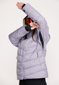 Lyslilla pufferjakke med quiltet design, lynlåslommer, justerbare manchetter og hætte, som en person med skibriller tager på.