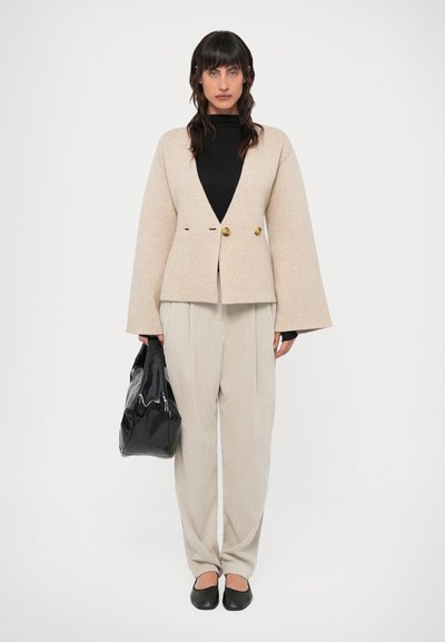 Blazer de mistura de lã bege com mangas largas e dois botões dourados, combinado com calças claras risca de giz e sapatos pretos, segurando uma bolsa preta.