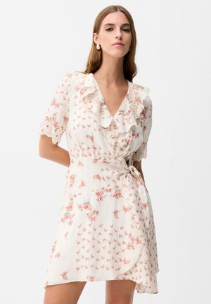 Femme portant une robe portefeuille blanche à imprimés floraux roses, avec un décolleté en V volanté et des manches courtes festonnées, sur un fond clair uni.