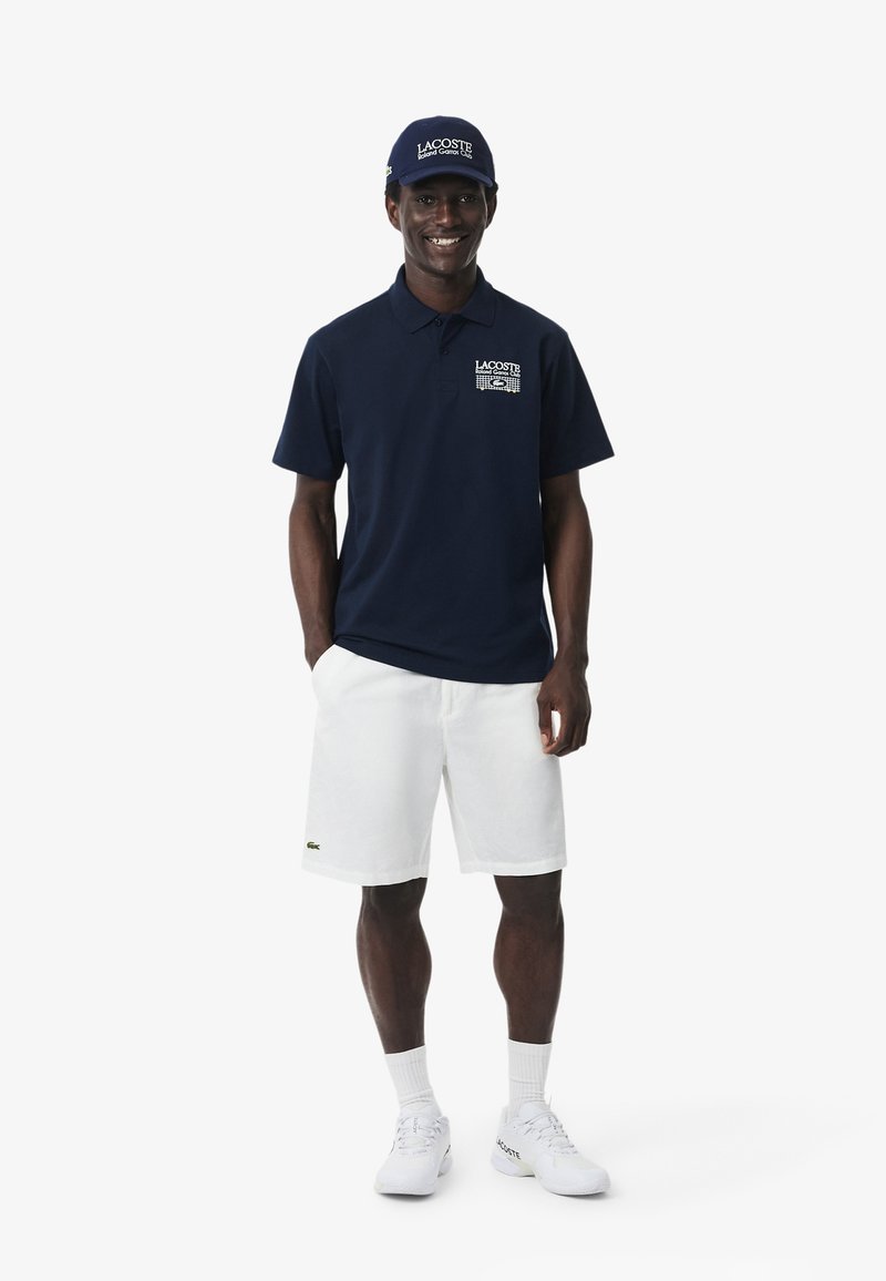 Homme portant un polo et une casquette Lacoste bleu marine avec un short blanc, des chaussettes et des baskets, debout les mains dans les poches, souriant.