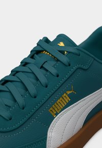 Δερμάτινο αθλητικό παπούτσι σε τυρκουάζ με λευκή πλαϊνή επιφάνεια, διακοσμημένο με χρυσό λογότυπο και κορδόνια. Η υφή της επιφάνειας και η λαστιχένια σόλα τονίζουν τον αθλητικό σχεδιασμό.