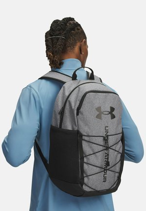 Sac à dos en tissu gris avec des accents noirs, sangles réglables, une poche en maille à l'avant et le logo Under Armour ; présente un détail de resserrement sur le devant.