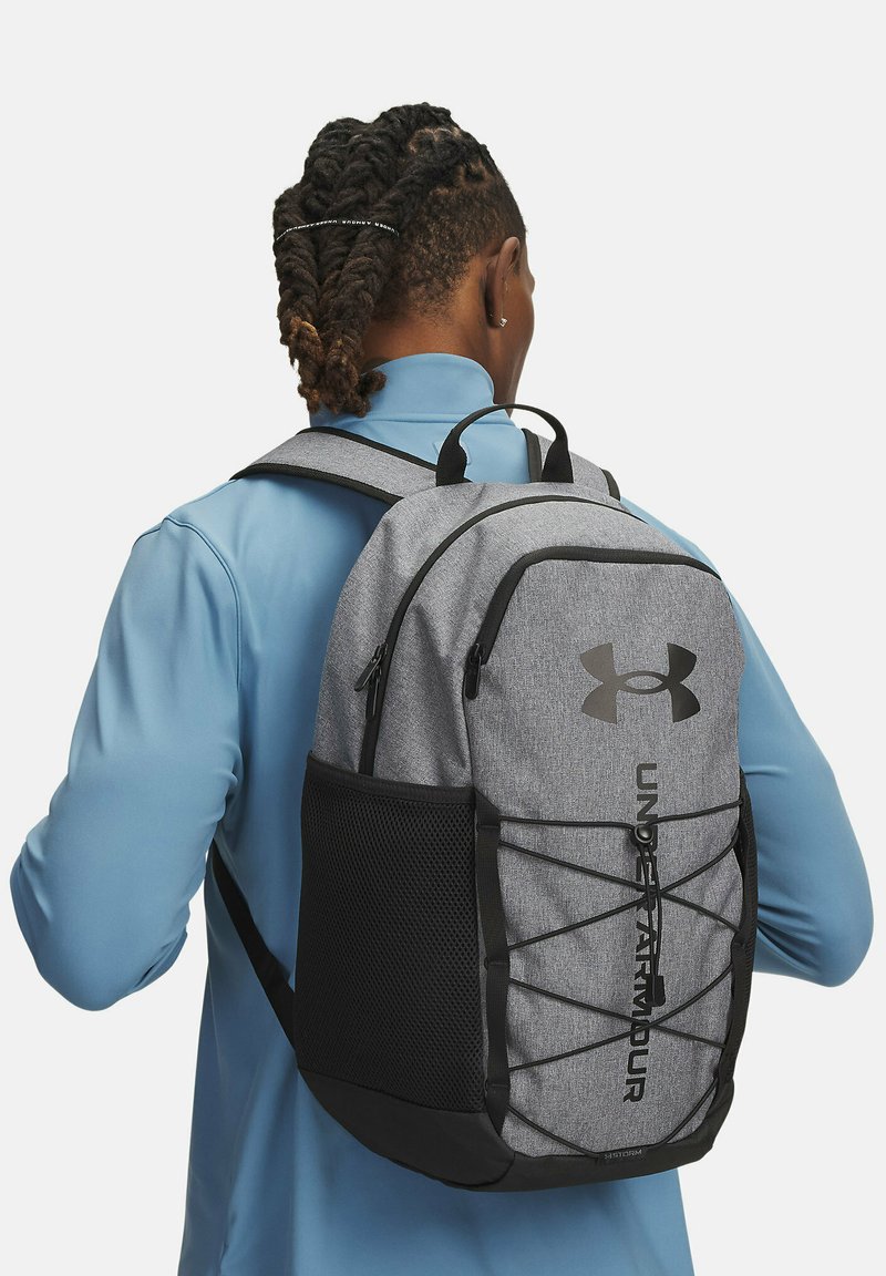 Zaino in tessuto grigio con accenti neri, spallacci regolabili, tasca anteriore in rete e logo Under Armour; presenta un dettaglio a coulisse sulla parte anteriore.