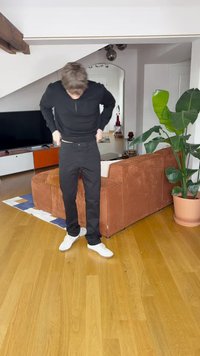 Svart skjorte med lange ermer og bukser med smal passform; hvite joggesko; teksturert brun sofa og potteplante i en moderne interiørsammenheng.