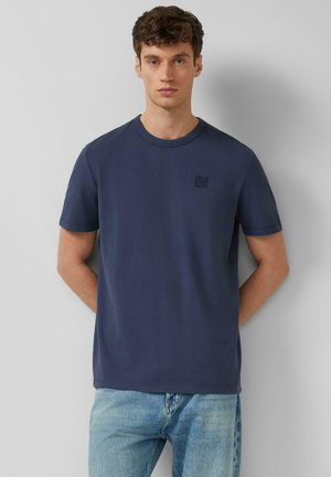 Jonge man met krullend bruin haar, gekleed in een effen marineblauw T-shirt en lichtblauwe jeans, staand tegen een lichtgrijze muur.