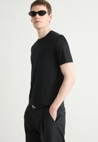 Maglietta nera a maniche corte in un tessuto liscio, abbinata a pantaloni neri e occhiali da sole neri a forma ovale, creando un look monocromatico coordinato.