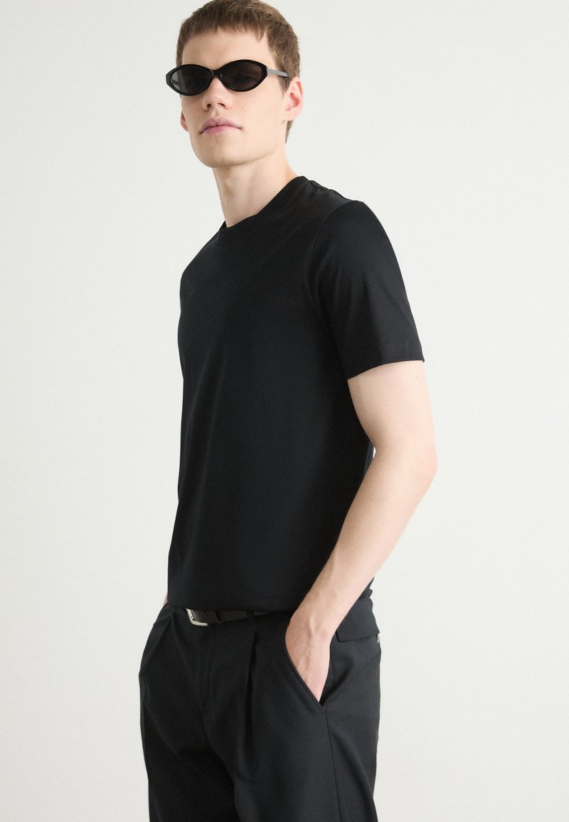 Maglietta nera a maniche corte in un tessuto liscio, abbinata a pantaloni neri e occhiali da sole neri a forma ovale, creando un look monocromatico coordinato.