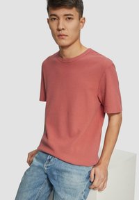 Korte mouwen, ronde hals T-shirt in stof in stof in stoffige roze met een gestructureerd oppervlak. Gecombineerd met lichtblauwe denim jeans. Tentoongesteld op een minimalistisch voetstuk.