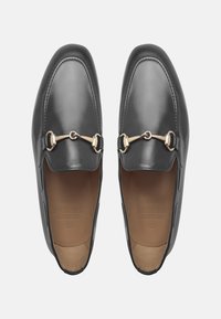 Mocassins en cuir noir avec une semelle plate, dotés d'un accent en mors en or et de détails cousus le long des bords. Texture lisse sur l'ensemble.