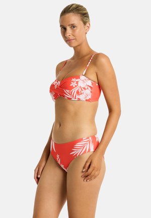 Kobieta w koralowym bikini z białym tropikalnym kwiatowym wzorem, stojąca bokiem na jednolitym jasnym tle.