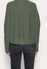 Grüner Strickpullover mit kastigem Schnitt, gerippter Textur und rundem Ausschnitt. Hat eine verkürzte Länge und überschnittene Schultern. Kombiniert mit dunkelblauen Jeans.