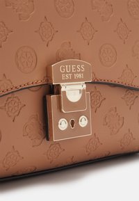 Hnědá texturovaná taška s kovovou zlatou sponou, na které je vyryto "GUESS EST 1981", s propracovanými vyraženými vzory a hladkým povrchem.