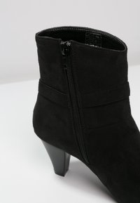 Botín de ante negro con cremallera lateral y tacón apilado de altura media sobre una superficie blanca.