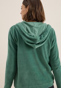 Femme vue de dos portant un sweat à capuche en tissu côtelé vert avec la capuche baissée, sur un fond clair uni.