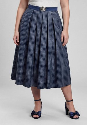 Femme portant une jupe midi plissée bleu marine à motif à carreaux, une ceinture décorative et des sandales à talons assorties avec des nœuds.