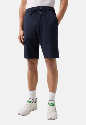 Mężczyzna ubrany w granatowe krótkie spodenki za kolano z regulowanym paskiem w talii i boczną kieszenią, białe skarpetki oraz białe sneakersy ze zielonymi sznurowadłami.