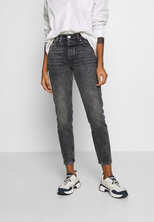 Person iført falmede sorte højtaljede jeans, hvid skjorte med opbage ærmer og hvide chunky sneakers med marineblå detaljer, stående på grå gulv.