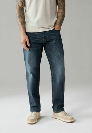Mørkeblå denimjeans med lige pasform, der har diskret slidmærker og en klassisk fem-lomme-design. Båret med beige sneakers.