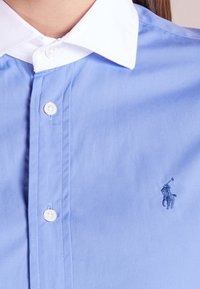 Blå button-up skjorta med vit krage och knappar, med en liten broderad polo-spelare-logotyp på bröstet.