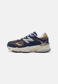 Tenisi din suede bleumarin și maro cu o talpă groasă crem, șireturi albe și accente din plasă. Prezintă un logo proeminent cu "N" pe lateral.