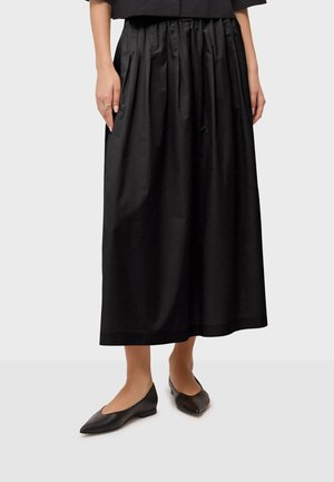 Donna che indossa una gonna midi nera plissettata abbinata a ballerine nere con punta appuntita, in piedi su uno sfondo chiaro semplice.