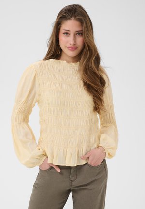 Jonge vrouw met lang golvend haar, gekleed in een lichtgele geplooide blouse en olijfgroene broek, staand met handen in de zakken tegen een witte achtergrond.