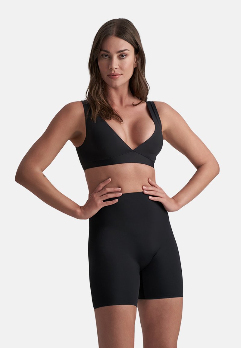 Donna con lunghi capelli castani, che indossa un reggiseno nero profondo a V e pantaloni corti neri a vita alta, in piedi con le mani sui fianchi davanti a uno sfondo bianco.